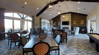This single-floor communal space exudes a refined, welcoming ambiance designed for both social gatherings and relaxed lounging. The room's most striking architectural feature is its soaring vaulted ceiling, accentuated by bold dark wood beams that add warmth and character to the open interior. Geometric pendant lanterns hang at varying heights, casting a soft, inviting glow throughout the space and complementing the room's upscale aesthetic.

Large arched and floor-to-ceiling windows line the walls, flooding the interior with generous natural light and offering views of the surrounding landscape. The dining area is furnished with dark wood rectangular tables paired with stylish upholstered chairs featuring a distinctive orange geometric pattern, adding a vibrant pop of color against the neutral palette.

Toward the rear of the space, a cozy seating area is anchored by a fireplace set into a richly textured dark accent wall, with a wall-mounted television above and flanking sconce lighting. Plush wing chairs and accent seating create an intimate gathering nook. The entire space is unified by a sophisticated patterned area rug in cool blue tones. Together, these elements create a polished, multi-functional environment ideal for dining, conversation, and relaxation.