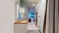This thoughtfully designed single-floor studio showcases a modern, minimalist aesthetic that maximizes every inch of available space. The open-plan layout seamlessly connects the living, sleeping, and kitchen areas, all unified by light hardwood flooring that extends throughout the main space.

The living area is anchored by a large window that allows natural light to fill the room, offering views of the surrounding building exterior. A built-in Murphy bed system is integrated into the wall, freeing up valuable floor space during the day. A compact sofa and a foldable wooden table complete the multifunctional living and sleeping zone.

The kitchenette is efficiently appointed with flat-front cabinetry in a soft grey tone, warm butcher block countertops, an undermount sink, a built-in induction cooktop, and an overhead microwave with range hood. Tall storage cabinets provide ample pantry and storage space alongside the entry door.

The bathroom transitions to grey tile flooring and features a streamlined vanity with a warm wood countertop, an undermount sink, and an illuminated mirrored medicine cabinet. A glass-enclosed shower and a private toilet area complete the well-organized bathroom. Frosted glass partitions and a curtain divider maintain privacy while preserving the open feel of the overall layout.