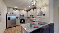 This single-floor residence offers a thoughtfully designed layout that balances comfort and functionality across its living spaces. The kitchen is a standout feature, showcasing white shaker-style cabinetry, granite countertops, stainless steel appliances, and a peninsula with seating — all illuminated by a modern globe pendant light and recessed lighting. Wood-look flooring flows seamlessly throughout the main areas, lending warmth and continuity to the space.

The open living and dining area is bright and welcoming, with natural light streaming through multiple windows dressed with layered curtains. A ceiling fan adds to the comfort of the living room, which offers ample space for seating and relaxation. The dining area features a charming round table accented by decorative arch-panel wall art, creating a cozy and stylish setting.

The bedrooms are carpeted for comfort and feature recessed lighting. One bedroom carries a playful, themed character, while another offers a more refined and serene atmosphere with tray ceiling details and soft neutral tones. The full bathroom includes a granite-topped vanity with gray cabinetry, a tub-shower combination, and wood-look flooring. A dedicated utility closet houses the water heater alongside wire shelving for additional storage.