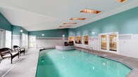 This single-level indoor aquatic facility offers a well-appointed environment designed for both recreation and relaxation. The centerpiece is a generously sized swimming pool with crystal-clear, blue-green water, accessible via stainless steel handrails and steps accented with decorative blue tile inlays. Adjacent to the main pool sits a rounded hot tub, seamlessly integrated into the pool deck and finished with matching tile detailing.

The architecture is defined by soaring vaulted ceilings that lend the space an open, airy quality. Multiple cedar-lined skylights are set into the ceiling, allowing natural light to filter in and brighten the interior. The walls feature a distinctive two-tone design, with teal paint above a wainscoting of clean white square tile, creating a fresh and cohesive aesthetic.

The pool deck is finished in a textured aggregate surface, providing both durability and slip resistance. Seating areas with wicker chairs are positioned near the windows, offering a comfortable spot to rest. Large windows along the perimeter frame views of the outdoors and allow additional natural light into the space. A pass-through window connects the pool area to an adjacent room, adding a convenient functional element. The overall ambiance is bright, welcoming, and well-maintained.