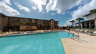 This outdoor pool area is a well-appointed amenity space designed for both relaxation and recreation. The centerpiece is a generously sized swimming pool with clear, blue-tinted water, accessible via stainless steel entry rails. Surrounding the pool is a broad, light-toned concrete sundeck furnished with rows of chaise lounges and outdoor dining sets, offering ample space for sunbathing and socializing under open skies.

Tall palm trees are strategically placed throughout the deck, adding a resort-like character to the setting. A multi-story building with a warm stucco and stone facade borders one side of the pool, providing an architectural backdrop. On the opposite side, a covered pavilion extends the usable outdoor space. This structure features prominent natural stone columns, wood-paneled ceilings, ceiling fans, and large tile flooring in a neutral palette—offering a shaded retreat from the sun. Glass railings along the pavilion maintain open sightlines to the pool area.

The design blends natural stone accents with contemporary finishes throughout, creating a cohesive and polished outdoor environment. The space benefits from abundant natural light and open sky views, contributing to an inviting and resort-inspired ambiance.