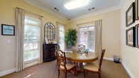 This dedicated closing room offers a polished and welcoming environment designed for professional consultations and lease signings. The space is anchored by a substantial round wooden table with a glass top, surrounded by ornate carved chairs with upholstered seats, providing a comfortable and formal setting for meetings. Warm yellow walls, accented by crisp white crown molding and baseboards, lend the room a classic, refined atmosphere. Natural light fills the space through multiple large windows dressed with elegant floor-length drapes and adjustable blinds, offering views of lush greenery outside. A dark carved sideboard along one wall adds a decorative touch, accompanied by an arched mirror that enhances the room's depth and traditional character. A tall decorative tree in the corner contributes a natural, welcoming element. The room is accessed through a glass-paned French door that connects to a broader hallway and adjacent common areas, with a framed gallery wall of architectural prints adding to the sophisticated décor. Neutral carpeting grounds the space, while a flush ceiling light ensures consistent, even illumination. Overall, this closing room balances professionalism with warmth, creating an inviting atmosphere for important transactions.