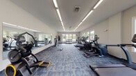 This fitness center is designed to support a comprehensive workout experience within a single, open-plan space. The room is finished with neutral white walls and patterned blue-grey carpet tiles, creating a clean and functional aesthetic throughout. Overhead, rows of linear LED lighting ensure the space is consistently and brightly illuminated.

Large windows line multiple walls, flooding the interior with natural light and offering views of the surrounding tree-lined landscape. A full-length mirror along one wall visually expands the space and supports strength training activities.

The cardio section is well-stocked with a variety of True-brand treadmills, elliptical trainers, stair climbers, and a stationary bike, arranged to allow comfortable spacing between machines. A rowing machine adds further variety to cardio options.

The strength training area includes a multi-use cable machine, a weight bench with barbell, and a tiered dumbbell rack offering a range of free weights. Functional training accessories such as stability balls and medicine balls are also available, providing options for flexibility and core-focused workouts.

The overall layout is organized and spacious, offering users an efficient and comfortable environment suitable for a wide range of fitness activities.