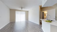 This single-floor space features an open-concept living area designed with a clean, functional layout. The living room is finished with wood-style laminate flooring in a neutral gray-brown tone that extends throughout, complementing the warm beige walls and white baseboards. A textured popcorn ceiling adds dimension overhead, while a ceiling fan with a built-in light fixture provides both airflow and ambient lighting.

A large sliding glass door dressed with vertical blinds serves as the focal point of the main living area, allowing an abundance of natural light to fill the room and providing access to an outdoor area. The open wall space offers flexibility for a variety of furniture arrangements.

A partial wall and raised countertop on the right side of the space suggests a pass-through or bar-style separation from an adjacent kitchen area, enhancing the open feel while defining distinct functional zones. A hallway visible in the background leads to additional rooms, hinting at a well-connected floor plan. Overall, the space presents a bright, neutral, and move-in-ready environment suitable for a range of living arrangements.