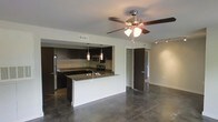 This single-floor space showcases a thoughtfully designed open-concept layout that seamlessly connects the kitchen and living areas. The polished concrete floors extend throughout, lending an industrial-modern aesthetic that anchors the overall design.

The kitchen is a standout feature, outfitted with dark espresso cabinetry that contrasts sharply against the white kitchen island, which is topped with granite countertops and equipped with a sink. Stainless steel appliances, including a refrigerator, microwave, and range, complement the space's modern character. Pendant lights hang above the island, adding focused illumination and a stylish touch.

The open living area benefits from a ceiling fan with integrated lighting, providing both comfort and practicality. A track lighting system is also present, offering flexible illumination options. The neutral white walls and open sightlines to adjacent rooms — including what appears to be a closet and additional room beyond — contribute to an airy, spacious feel. The combination of industrial flooring, dark finishes, and contemporary fixtures gives this space a clean, urban character.