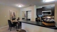 This single-floor residence showcases an open-concept layout that seamlessly connects the kitchen and dining areas, creating a cohesive and functional living environment. The kitchen is outfitted with dark espresso cabinetry that extends to the upper walls, providing ample storage space. A dark granite-style countertop extends into a peninsula that serves as both a workspace and a casual bar-style seating area, effectively defining the boundary between the kitchen and the adjoining dining space. Stainless steel appliances, including a microwave and range, complement the kitchen's contemporary aesthetic, while a stylish arc faucet adds a refined touch. A tile backsplash adds texture behind the cooking area. The dining space accommodates a full table and chairs, decorated with framed abstract artwork on the walls, contributing a warm and inviting atmosphere. A built-in shelving unit is visible in the background, offering additional display or storage options. The textured ceiling and flush-mount light fixture provide consistent illumination throughout the open space, while neutral wall tones tie the entire area together cohesively.