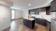 This single-floor residence showcases a thoughtfully designed open layout with rich, dark hardwood-style flooring running throughout. The kitchen serves as a functional centerpiece, featuring dark espresso cabinetry extending to the ceiling, white quartz countertops, a subway tile backsplash, and a central island with built-in wine storage. Stainless steel appliances — including a gas range, microwave, refrigerator, and dishwasher — complete the space. A ceiling fan is positioned in the adjoining living area, which receives natural light through large windows fitted with horizontal blinds.

A hallway connects the main living area to the private rooms, leading to a bedroom that features its own ceiling fan, large window, and access to a dedicated walk-in closet with a full rod and shelving system. The bathroom includes a full soaking tub and shower combination with chrome fixtures and a sleek glass partition. A separate utility closet houses a stacked washer and dryer unit, providing convenient in-unit laundry. The overall design is clean and contemporary, with neutral white walls and gray trim framing the space throughout.