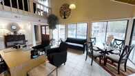 This single-floor common area is designed to be both functional and inviting, offering a bright and open atmosphere that encourages relaxation and social interaction. The space is anchored by impressively high ceilings that create a sense of grandeur, complemented by an upper-level overlook that adds architectural depth and visual interest. Large, floor-to-ceiling windows flood the interior with natural light and frame views of an outdoor pool area, seamlessly connecting the indoors with the surrounding environment. The main seating area features a set of sleek black upholstered chairs and a loveseat arranged for easy conversation, alongside a dining table with traditional wooden chairs for more structured gatherings. Decorative touches, including a bold metal tree wall art piece and a lush indoor tree, add warmth and character to the neutral-toned walls and light tile flooring. A classic wooden French door with glass panels provides an entryway that enhances the welcoming feel of the space. The overall ambiance is clean, modern yet comfortable, making this a versatile gathering space suited for both casual lounging and community activities.