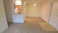 This single-floor residential space offers a practical and comfortable layout designed for everyday living. The main living area is generously sized, featuring wall-to-wall carpeting in a neutral beige tone that adds warmth underfoot. A ceiling fan with an integrated light fixture is centrally positioned, providing both air circulation and ambient lighting throughout the space. The light-colored walls contribute to a bright, airy atmosphere.

The kitchen is connected to the living area via a pass-through counter with a dark stone or laminate surface, creating an open, communicative feel between the two spaces. Warm wood-toned cabinetry lines the upper and lower sections of the kitchen, offering ample storage. A white refrigerator is visible, complementing the clean, functional aesthetic of the kitchen. The pass-through design allows for easy interaction between the kitchen and living room, making the space well-suited for both daily routines and casual entertaining.

Overall, the space presents a straightforward and functional interior with a cohesive, neutral color palette. The open connection between the living and kitchen areas enhances the sense of space, making this a practical and welcoming single-floor residence.