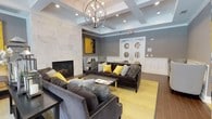 This community room is a thoughtfully designed gathering space that blends elegance with comfort. The architectural centerpiece is a coffered ceiling adorned with a striking orb chandelier, which casts warm light throughout the room and enhances the sense of height and openness. Recessed lighting further brightens the space, complementing the natural palette of cool greys and crisp whites.

A floor-to-ceiling marble-surround fireplace serves as the room's focal anchor, adding a refined, sophisticated touch. Flanking the fireplace are bold yellow abstract artworks that inject energy and personality into the space. The seating arrangement features plush, charcoal-grey velvet sofas and accent chairs, accented with yellow and textured throw pillows, all centered around a sleek coffee table atop a vivid yellow area rug.

White wainscoting lines the lower walls, providing classic architectural detail that contrasts beautifully with the grey upper walls. A console table styled with lanterns, candles, and fresh florals adds a decorative layer of warmth. Double French doors with circular glass accents at the rear of the room offer an elegant entry point, completing this inviting and well-appointed community space.