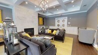 This community room is a thoughtfully designed gathering space that blends elegance with comfort. The architectural centerpiece is a coffered ceiling adorned with a striking orb chandelier, which casts warm light throughout the room and enhances the sense of height and openness. Recessed lighting further brightens the space, complementing the natural palette of cool greys and crisp whites.

A floor-to-ceiling marble-surround fireplace serves as the room's focal anchor, adding a refined, sophisticated touch. Flanking the fireplace are bold yellow abstract artworks that inject energy and personality into the space. The seating arrangement features plush, charcoal-grey velvet sofas and accent chairs, accented with yellow and textured throw pillows, all centered around a sleek coffee table atop a vivid yellow area rug.

White wainscoting lines the lower walls, providing classic architectural detail that contrasts beautifully with the grey upper walls. A console table styled with lanterns, candles, and fresh florals adds a decorative layer of warmth. Double French doors with circular glass accents at the rear of the room offer an elegant entry point, completing this inviting and well-appointed community space.