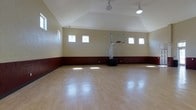 This single-story indoor basketball court offers a spacious and well-appointed environment designed for athletic activity. The vaulted ceiling rises to a generous apex, creating an open, airy atmosphere with ample vertical clearance for play. A regulation-height adjustable basketball hoop with a clear backboard and a portable base is positioned at one end of the court, ready for use.

The flooring consists of smooth, light-toned hardwood-style planks that reflect natural and artificial light throughout the space, contributing to its bright and inviting feel. The walls feature a two-tone treatment, with a deep burgundy wainscoting along the lower portion and a warm cream finish above, providing a classic gymnasium aesthetic.

Natural light filters in through a series of evenly spaced, small rectangular windows positioned high along both side walls, supplemented by industrial-style pendant light fixtures hanging from the ceiling peak. The combination of natural and artificial lighting ensures consistent illumination across the entire court. Overall, the space is clean, functional, and well-suited for both competitive play and casual recreation.