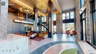 Located in Morrisville, NC, this dynamic two-story amenity space makes a bold architectural statement from the moment you enter. The ground floor welcomes visitors with a soaring double-height lobby anchored by striking stacked-stone accent columns, large floor-to-ceiling windows that flood the space with natural light, and sweeping outdoor views. A sleek marble reception desk anchors the entry, while a colorful curved carpet design flows across the polished tile floor beneath dramatic sputnik chandelier lighting. Eclectic lounge seating, vibrant area rugs, and arcade game stations give the space a lively, social energy. A sculptural open-tread staircase with wood and steel detailing connects the two floors, complemented by a built-in bench seating nook beneath it. The upper level continues the bold design language with herringbone wood flooring, a glass-enclosed office suite featuring a black marble conference table, pendant lighting, and private individual workstations brightened by cobalt blue accent walls and colorful artwork. A cozy reading nook with a bookshelf and plush seating rounds out the upper level. Throughout both floors, the design blends industrial textures with vibrant contemporary furnishings, creating an engaging and highly functional communal environment.