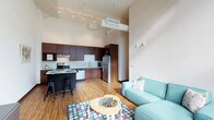 This single-floor space showcases a thoughtfully designed open-concept layout that seamlessly blends the kitchen and living areas into one cohesive, functional environment. Soaring ceilings amplify the sense of space and airiness, while warm hardwood-style flooring runs throughout, adding a welcoming touch to the modern aesthetic.

The kitchen is well-appointed with rich dark wood cabinetry, stainless steel appliances including a refrigerator and range, a double sink, and a breakfast bar with seating — ideal for casual dining or entertaining. A track lighting system overhead provides both task and ambient illumination, complementing the natural light that fills the space.

The living area flows naturally from the kitchen, offering a comfortable and stylish setting anchored by a teal sofa, a round accent table, and a colorful area rug that adds personality to the space. Exposed ductwork and industrial-style ceiling details lend the unit a contemporary loft-like character, balancing modern design with functional living. The overall ambiance is clean, bright, and inviting, making it a versatile space suited for both relaxing and everyday living.