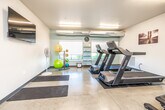 This fitness center is a fully equipped, single-floor exercise facility designed to support a wide range of workout routines. The space features polished concrete flooring throughout, with dedicated rubber mat zones for both cardio equipment and floor exercises. Overhead fluorescent strip lighting ensures the entire area is brightly and evenly illuminated, complemented by natural light streaming in through multiple windows fitted with white horizontal blinds, offering views of surrounding greenery.

The cardio zone is anchored by two Vision Fitness treadmills positioned side by side, alongside a flat utility bench. A dedicated strength training area includes a multi-function cable machine and a dumbbell rack stocked with a full range of free weights, accompanied by an adjustable weight bench. For functional and flexibility training, the space provides an assortment of stability balls, medicine balls, and several exercise mats laid out on the floor.

A wall-mounted flat-screen television adds an entertainment element, while a standing fan supports air circulation alongside exposed HVAC ductwork running along the ceiling. Open shelving near the windows organizes smaller accessories and equipment. The space is clean, practical, and efficiently arranged to accommodate multiple users simultaneously across its distinct training zones.