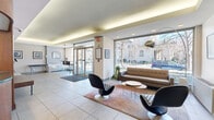 Located at 1616 18th St NW in Washington, DC's vibrant Dupont Circle neighborhood, this building lobby offers a well-appointed and functional entry experience. The space is designed with a contemporary aesthetic, featuring large floor-to-ceiling windows along the street-facing wall that flood the interior with abundant natural light and provide open views of the tree-lined surroundings outside.

The main lounge area is furnished with mid-century modern-style seating, including a sleek upholstered sofa, sculptural pod-style chairs, and a set of round accent tables arranged atop a warm area rug — creating an inviting atmosphere for residents and guests. A wall-mounted flat-screen display and a wooden console table with a water cooler add practical amenities to the sitting area.

A standout feature is the curved, warm wood reception desk, which anchors the rear of the lobby with an elegant architectural presence. Rich wood paneling lines the surrounding walls, offering a refined contrast to the light-toned tile flooring that extends throughout the space. Framed artwork decorates the walls, contributing a polished, curated feel.

A set of tiled steps leads to a staircase providing access to upper floors, while recessed and cove lighting along the ceiling enhances the lobby's clean, modern ambiance. The entrance is defined by glass double doors with a commercial-grade entry mat, completing this welcoming and professionally designed lobby space.