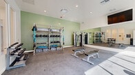 This modern fitness center in Lathrop, California is thoughtfully designed to support a full range of workout routines. The space is divided into two distinct zones, each tailored to a specific type of training. The cardio area is bright and airy, featuring large floor-to-ceiling windows that flood the room with natural light and offer views of an outdoor pool. This section is outfitted with treadmills, ellipticals, and stationary cycling bikes, alongside a tiered dumbbell rack for free weight exercises. A water station and restroom facilities are conveniently accessible from this area, along with a circular piece of contemporary wall art that adds a touch of character. The strength and functional training studio is finished with sage green accent walls and carpeted flooring, giving it a distinct, energetic atmosphere. It is equipped with a RaX multi-functional training rack loaded with medicine balls, resistance bands, and TRX suspension straps, as well as a cable machine and adjustable workout benches. A large wall-mounted mirror enhances the sense of space and supports form-checking during exercises. A mounted flat-screen TV is positioned for easy viewing throughout the room. Recessed lighting and high ceilings complete the clean, professional aesthetic of this comprehensive fitness facility.