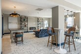 Located at 55 Park Ave in Bay Shore, NY, this thoughtfully designed single-floor lounge space offers a refined yet welcoming atmosphere with a cohesive eclectic aesthetic. The open layout is anchored by a generously sized seating area featuring a deep navy velvet sofa accented with teal and coral throw pillows, paired with richly upholstered multicolored armchairs and a tufted cognac leather ottoman set atop a textured area rug. A sleek wall-mounted linear fireplace adds warmth and visual interest, flanked by an industrial-style open shelving unit displaying curated décor.

Natural light pours in through multiple large windows positioned throughout the space, offering sweeping views of the surrounding Bay Shore skyline and creating bright, sun-drenched seating nooks. Pendant lighting in bold geometric forms adds character overhead. A high-top bar area with sculptural tulip-style tables and walnut-and-navy barstools provides an additional casual gathering spot with window views.

The kitchenette is a standout feature, clad in a striking iridescent teal subway tile backsplash complemented by warm wood cabinetry, a compact refrigerator, microwave, and utility sink — ideal for hosting events. A full-length mirror and additional lounge chairs complete the space, lending it an inviting, versatile character.