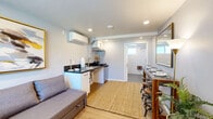Located at 530 W Wilson St in Costa Mesa, California, this thoughtfully designed single-floor studio unit makes efficient use of its compact footprint. The open-plan living and kitchen area is bright and welcoming, featuring light wood-look flooring, recessed lighting, and a wall-mounted mini-split HVAC system for year-round climate control. A sectional sofa serves as the centerpiece of the living space, complemented by a woven area rug and a window that allows natural light to filter in. A framed abstract artwork adds a bold decorative accent to the neutral-toned walls. The kitchenette is neatly integrated along one wall, offering white cabinetry, a dark granite-style countertop, a single-basin sink, and space for a small dining bar with stools — maximizing functionality without sacrificing style. The bathroom is a standout feature, designed with full accessibility in mind. It includes a spacious open wet room with textured wave-pattern wall tiles, grey mosaic floor tiles, a rain showerhead, a handheld shower fixture, grab bars, a fold-down shower bench, and a pedestal sink beneath a gold-framed mirror. Natural light enters through a frosted window, keeping the space bright and airy. This unit delivers a clean, modern aesthetic suited for comfortable everyday living.