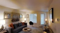 This single-floor residence offers a thoughtfully designed living area that balances comfort and contemporary style. The living room serves as the central gathering space, characterized by neutral beige walls and carpeted flooring that contribute to a warm, inviting ambiance. A large window fitted with horizontal blinds spans a corner of the room, allowing generous amounts of natural light to fill the space while also providing access to what appears to be an exterior door, suggesting a patio or outdoor area beyond.

The room is styled with a dark charcoal sofa accented by patterned and rust-colored throw pillows, paired with a sleek Eames-style accent chair that adds a modern touch. A round, glass-top coffee table sits at the center, contributing to an open and airy feel. Wall art, including a large landscape canvas, adds visual depth and character to the space.

Ambient lighting is provided through strategically placed floor and table lamps, creating a soft, layered glow that complements the natural light. The overall aesthetic is modern yet relaxed, making the living area a functional and visually appealing space for everyday living and entertaining.