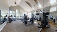 Located at 5400 Chenonceau Blvd in Little Rock, Arkansas, this single-floor fitness center is designed to support a comprehensive workout experience. The space is defined by its dramatic vaulted ceilings, which lend an open, airy quality to the room and are complemented by a combination of recessed lighting, pendant fixtures, and ceiling fans. Large multi-pane windows line the walls, flooding the interior with natural light and offering views of the surrounding greenery and community exterior.

The main workout area is generously proportioned and equipped with a wide variety of strength-training machines, including cable systems, leg press units, and multi-function weight stations. A full dumbbell rack is neatly organized along one mirrored wall, which further enhances the sense of openness and allows for easy monitoring of form. Cardio equipment, including treadmills with individual display screens, is positioned along the windowed perimeter to take advantage of the outdoor views. Wall-mounted televisions provide additional entertainment options throughout the space.

A compact kitchenette area is also included, featuring rich dark wood cabinetry, granite countertops, a stainless steel sink, and a dishwasher — offering convenient amenities for users of the fitness center. The overall ambiance is clean, modern, and well-maintained, making this facility a functional and inviting space for daily exercise.