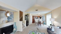 This two-story home showcases an open, airy layout anchored by a striking vaulted ceiling in the main living area, which adds a strong sense of vertical space and grandeur. The living room flows seamlessly into the dining area, creating an interconnected space ideal for both everyday living and entertaining. Plush wall-to-wall carpeting in a warm neutral tone runs throughout, contributing to the home's comfortable and inviting atmosphere.

The dining area accommodates a glass-top table with upholstered seating, positioned adjacent to the kitchen. The kitchen features warm wood cabinetry, light countertops, and built-in appliances, offering a functional and cohesive design. A pass-through or open connection between the kitchen and dining space enhances the home's open-concept feel.

A staircase with white railings leads to the second floor, adding architectural interest and defining the transition between levels. The walls are painted in soft, warm tones that complement the natural light filtering through the windows. A display cabinet near the staircase provides both storage and a decorative focal point, rounding out the well-appointed main level.