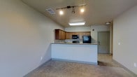 This single-floor space showcases an open and functional layout that blends the kitchen and living areas into one cohesive environment. The kitchen is a standout feature, equipped with warm-toned wood cabinetry, granite countertops, and black appliances including a refrigerator, microwave, and range. A peninsula-style counter extends outward, creating a natural divide between the kitchen and the adjacent living area while also offering additional workspace or casual dining space. Track lighting mounted on the ceiling provides focused illumination throughout the kitchen zone, complementing the flush-mount ceiling light nearby. The living area features neutral-toned carpet flooring, contributing to a comfortable and inviting atmosphere. Walls are finished in a soft, neutral tone that enhances the overall brightness of the space. Ceiling vents are strategically placed for efficient climate control throughout the unit. A paneled entry door is visible in the background, offering a clear sense of the layout's flow. The space presents a clean, move-in ready aesthetic with practical design choices that balance form and function effectively.