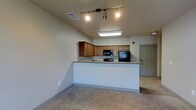 This single-floor space showcases an open and functional layout that blends the kitchen and living areas into one cohesive environment. The kitchen is a standout feature, equipped with warm-toned wood cabinetry, granite countertops, and black appliances including a refrigerator, microwave, and range. A peninsula-style counter extends outward, creating a natural divide between the kitchen and the adjacent living area while also offering additional workspace or casual dining space. Track lighting mounted on the ceiling provides focused illumination throughout the kitchen zone, complementing the flush-mount ceiling light nearby. The living area features neutral-toned carpet flooring, contributing to a comfortable and inviting atmosphere. Walls are finished in a soft, neutral tone that enhances the overall brightness of the space. Ceiling vents are strategically placed for efficient climate control throughout the unit. A paneled entry door is visible in the background, offering a clear sense of the layout's flow. The space presents a clean, move-in ready aesthetic with practical design choices that balance form and function effectively.