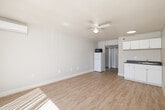 Located at 17065 West Bernardo Drive in San Diego, CA, this efficiently designed single-floor unit offers a well-organized layout comprising a living room, kitchenette, bathroom, closet, hallway, and patio. The living area is bright and open, featuring light wood-tone laminate flooring, a ceiling fan with integrated lighting, and a wall-mounted mini-split air conditioning unit for climate control. A large sliding glass door floods the space with natural light and provides direct access to the patio, offering views of the surrounding outdoor area. The kitchenette is neatly appointed with white cabinetry, a dark countertop, a single-basin sink, and a refrigerator, making efficient use of the available space. The bathroom presents a clean, refined aesthetic with light tile flooring, a granite-topped vanity, a large framed mirror, and glass shelving, lending a touch of elegance to the compact room. A dedicated closet and hallway provide practical storage and circulation between spaces. The unit's crisp white walls and abundant natural light contribute to an airy, functional ambiance throughout.