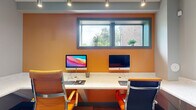 This business center is a compact yet efficiently organized workspace designed to meet professional productivity needs. The room is entered through a distinctive glass-paneled door with frosted privacy banding, labeled "Business Center," giving it a polished, professional identity from the moment you arrive.

Inside, the space is divided into distinct functional zones. A bold accent wall in warm amber-orange anchors the primary workstation area, where a built-in, white terrazzo-style countertop runs along the wall and accommodates two iMac desktop computers side by side, each paired with an ergonomic orange leather task chair. A wide, dark-framed horizontal window above the desk allows natural light to filter in and offers a view of greenery outside, with small potted plants lining the sill.

A separate alcove provides a quieter, individual workstation featuring an additional iMac and matching orange chair, set against neutral gray walls and accented with decorative wall art.

The printing and copying station is positioned near the entry door, offering a multifunction printer on a continuous countertop surface alongside a sleek stainless steel waste bin. A framed abstract artwork in gold and earth tones adds a decorative touch to this area.

Wood-look luxury vinyl plank flooring in mixed gray and brown tones flows throughout, complemented by recessed and track lighting overhead that ensures consistent, well-distributed illumination across the entire space.