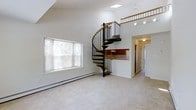 Located at 1202 Crescent Dr in Tarrytown, NY, this one-bedroom, one-bathroom loft offers a distinctive two-story layout that maximizes both space and architectural character. The main living area immediately impresses with soaring ceilings that create an airy, open atmosphere, enhanced by a skylight that floods the space with natural light. Soft carpeting extends throughout the living area, contributing to a warm and comfortable environment. A striking spiral staircase with a wood-and-metal design serves as the visual centerpiece of the main floor, connecting the living level to the upper loft. Large windows with blinds frame views of the surrounding greenery, bringing the outdoors in and adding brightness to the space. Adjacent to the living area, a compact kitchen features warm wood cabinetry and essential appliances, with a pass-through that maintains the open feel. A hallway leads to the bathroom and additional storage. The upper loft level, accessible via the spiral staircase, is framed by a wood railing and provides versatile space ideal for a bedroom or home office. Track lighting on the upper level ensures functional illumination throughout. This thoughtfully designed loft combines charm, functionality, and distinctive architectural details in the heart of Tarrytown.