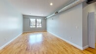 Located at 26 Portland St in Worcester, MA, this single-floor unit offers a well-designed open layout with consistent hardwood flooring throughout. The main living area is generously proportioned, featuring light gray walls, recessed lighting, and a large window that frames views of the surrounding cityscape, allowing natural light to fill the room. An exposed silver ventilation duct runs along the upper wall, adding an industrial accent to the otherwise clean and modern aesthetic.

The kitchen is positioned along one wall in an efficient galley-style configuration. It is equipped with warm-toned wood cabinetry, granite countertops, a stainless steel range with built-in microwave, and a full-size refrigerator. The kitchen flows seamlessly into the main living space, making it practical for both everyday use and entertaining.

The bathroom is accessed through a paneled white door and features tile flooring in a neutral tone, a full-size bathtub with overhead shower, and white fixtures throughout, presenting a clean and functional design.

Overall, the unit maintains a cohesive, contemporary feel with its neutral gray palette, polished hardwood floors, and practical layout — offering a comfortable and functional living environment in the heart of Worcester.