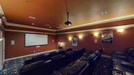 This purpose-built media room in Spring, TX, is designed to deliver an authentic cinematic experience in a private setting. The room is finished in deep, rich terracotta-red walls accented by cream crown molding, creating a warm and immersive theater ambiance. Recessed ceiling lights and softly glowing wall sconces provide carefully controlled ambient lighting that enhances the movie-watching atmosphere without compromising screen visibility.

At the front of the room, a large projection screen spans the wall, paired with a ceiling-mounted projector for a true big-screen experience. Seating is arranged in tiered rows of plush, black leather recliner chairs equipped with built-in cup holders, ensuring comfort for all viewers regardless of their position. A classic-style popcorn machine stands near the front, adding a charming, authentic movie theater touch.

The flooring features a bold, decorative patterned carpet with colorful motifs reminiscent of classic cinema carpeting, further reinforcing the theatrical theme. Framed movie posters adorn the walls, contributing to the room's cinematic character. The space includes a clearly marked exit door and a secondary entry point, ensuring practical accessibility. Crown molding details along the ceiling add an elegant finishing touch, making this media room as refined as it is functional.