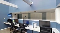 This single-floor space functions as a dedicated business center, designed to provide a functional and comfortable environment for work and connectivity. The room is characterized by its notably high ceilings, which lend an open, airy quality to the space and contribute to an overall sense of spaciousness. The walls are painted in a cool, calming blue tone that runs consistently throughout, creating a cohesive and professional atmosphere.

A continuous white workstation counter spans the length of the room, accommodating multiple desktop computer setups — each equipped with a full-sized monitor, keyboard, and mouse. Ergonomic mesh-back office chairs are positioned at each station, supporting extended periods of use. Framed informational signage is placed along the counter, offering guidance or notices to users.

Recessed ceiling lighting provides even illumination across the workspace. Along the back wall, louvered cabinet panels offer a clean, organized appearance while concealing storage or utility infrastructure. An accessible restroom is conveniently located nearby, as indicated by visible signage. Security is supported by a ceiling-mounted camera, ensuring a safe environment for all users.