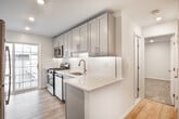 Located at 58 Piedmont Dr in Port Jefferson Station, NY, this thoughtfully updated single-floor unit showcases a clean, modern aesthetic throughout. The kitchen is a standout feature, equipped with sleek gray shaker-style cabinetry that extends to the ceiling for ample storage, white quartz countertops with a peninsula, a classic white subway tile backsplash, a brushed gold faucet, and stainless steel appliances including a gas range with microwave above. Light wood-style flooring runs through the main living areas, complementing the bright, neutral palette.

The open living and dining area benefits from recessed lighting and a pass-through detail that connects seamlessly to the kitchen, creating an inviting and functional layout. A separate room with the same light flooring and baseboard heating provides flexible living space with natural light from a large window overlooking a residential neighborhood.

The bedrooms feature plush carpeting, recessed lighting, wall-mounted climate control units, and baseboard heating. One bedroom includes generous built-in closet space with dual rods and shelving.

The full bathroom is finished with neutral tile flooring, a modern wood-toned vanity with a white countertop, brushed gold fixtures, and a tub-shower combination with a window for natural ventilation. Throughout the unit, clean white walls and consistent finishes create a cohesive, move-in-ready environment.