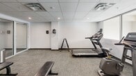 Located at 1355 Commissioners Rd W in London, Ontario, this fitness facility offers a well-organized and efficiently designed workout space. The gym features a drop ceiling with recessed lighting and integrated ventilation, creating a bright and comfortable environment. Flooring throughout is a durable, low-pile carpet tile in a neutral grey tone, well-suited for a fitness setting.

The space is equipped with a variety of cardio machines, including a treadmill, upright stationary bike, and an elliptical trainer. A dedicated free weights area features a tiered dumbbell rack with multiple weight increments, accompanied by flat workout benches for strength training exercises. Wall-mounted sanitation dispensers are conveniently positioned for users.

Natural light enters through windows along one wall, offering a view to the exterior. A striking geometric chandelier accent near the window wall adds an unexpected decorative touch to the otherwise functional space. A glass partition wall with a full-length glass door separates the main gym area from a small anteroom, which includes a desk and waste receptacle — ideal for sign-in or equipment management purposes.

Overall, the gym presents a clean, modern aesthetic with a practical layout that maximizes the use of the available space for a range of fitness activities.