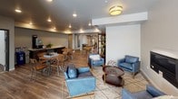 This well-appointed single-floor community space is designed to foster comfort and social interaction across several distinct yet open zones. The entry lobby sets a welcoming tone with warm wood-look flooring, recessed lighting, and a striking round table centerpiece set atop a cowhide rug, flanked by multiple access doors that allow for easy flow throughout the facility. Adjacent to the lobby, a spacious lounge area offers a relaxed gathering environment with contemporary blue leather armchairs, a patterned area rug, and a sleek wall-mounted fireplace that serves as a stylish focal point. The open layout connects seamlessly to a casual dining and game area furnished with round wood-top tables and metal chairs, where a chess set adds an inviting recreational touch. A built-in beverage station along one wall features dark wood cabinetry, a countertop coffee machine setup, and decorative greenery, providing residents or guests with convenient refreshment options. Throughout the space, consistent wood-look flooring, neutral gray walls, and a combination of recessed and flush-mount ceiling lighting create a cohesive, modern aesthetic. The thoughtful arrangement of zones makes this community space equally suited for quiet relaxation, social gatherings, and casual recreation.