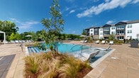 Located at 2035 Tapestry Park Drive in Indian Land, South Carolina, this outdoor amenity space is a thoughtfully designed retreat that serves the surrounding multi-story residential community. At its centerpiece is a generously sized resort-style swimming pool with crystal-clear blue water, complemented by in-pool lounge chairs that allow residents to relax directly in the water. The expansive stone-paved sun deck surrounds the pool and provides ample space for additional chaise lounges and shaded umbrella seating, ideal for soaking up the South Carolina sunshine. A sleek white pergola anchors one section of the deck, offering a shaded outdoor living area furnished with contemporary sofas and accent chairs — perfect for socializing or unwinding. The grounds are accented with ornamental grasses, young trees, and carefully curated landscaping beds that add texture and natural warmth to the hardscape. A gated entry with a paver walkway provides secure access to the amenity area. Black iron fencing encloses the space, lending a clean, upscale aesthetic while maintaining open sightlines to the surrounding greenery and the architecturally distinct multi-story buildings featuring dark siding, white trim, and balconies that frame the backdrop of the outdoor oasis.