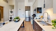 Located at 1600 Broadway in Sacramento's 95818 neighborhood, this well-appointed one-bedroom, one-bathroom unit offers a thoughtfully designed open-concept layout across a single floor. The spacious bedroom serves as the heart of the unit, featuring soaring ceilings, a ceiling fan, and floor-to-ceiling windows that flood the room with natural light and frame views of the surrounding streetscape and mature trees. Light-toned hardwood flooring flows seamlessly throughout, enhancing the bright, airy ambiance.

The kitchen is outfitted with modern two-tone cabinetry—combining rich dark wood lower cabinets with light upper cabinets—paired with a white tile backsplash, stainless steel appliances including a refrigerator, range, and microwave, and a sleek light-colored countertop island that doubles as a dining surface. Pendant lighting adds a contemporary finishing touch.

A stacked washer and dryer unit is conveniently integrated into the layout near the kitchen area. The bathroom features tile flooring, a vanity with a backlit mirror, and a full tub-shower combination with tile surround. Clean white walls and recessed lighting throughout lend the entire space a polished, modern aesthetic.