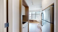 This thoughtfully designed urban unit occupies a single floor and makes excellent use of every square foot through smart, built-in features and a cohesive modern aesthetic. Warm wood-toned cabinetry and light hardwood flooring run throughout, creating a welcoming contrast against the crisp white walls and ceilings fitted with track lighting.

The main living area is anchored by a large picture window that frames sweeping city skyline views, flooding the space with natural light. A built-in upholstered window seat with open shelving below doubles as both a relaxation nook and practical storage. A compact dining area with a round white table and contemporary chairs sits adjacent, alongside a wall-mounted flat-screen television.

The kitchenette is efficiently organized, featuring light wood cabinetry, a bold teal glass backsplash, white countertops, a built-in cooktop, over-the-range microwave, and a full-size stainless steel refrigerator. A custom wood-panel divider elegantly separates the kitchen from the sleeping area.

The sleeping area accommodates a platform bed with integrated drawer storage beneath, maximizing functionality. A mirrored panel and a built-in bedside unit complete the space.

The bathroom maintains the clean, modern palette with large-format tile flooring, a floating wood-front vanity, and a walk-in shower, offering a refined and practical finish to this well-appointed urban retreat.