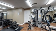 This single-floor fitness room in Laurel is designed to provide a practical and well-equipped exercise environment. The space features warm, neutral-toned walls complemented by wood-look flooring, creating a clean and functional atmosphere. Overhead fluorescent lighting ensures the room is consistently and evenly illuminated throughout.

The centerpiece of the room is a multi-station cable and weight machine, offering a wide range of strength-training exercises suitable for various fitness levels. An adjustable workout bench and a stability ball provide additional options for resistance and core training. An exercise mat on the floor offers a dedicated area for floor-based workouts. Instructional fitness charts are mounted on the walls, serving as convenient reference guides during training sessions.

Full-length mirrors line one wall, enhancing the sense of space and allowing users to monitor their form while exercising. A white door provides access to an adjoining hallway, contributing to the room's clean, organized appearance. The textured ceiling and visible HVAC vents reflect a practical, no-frills design focused on functionality, making this fitness room a straightforward and efficient space for regular exercise.