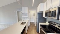 This single-floor residence showcases a thoughtful layout with clean, contemporary finishes throughout. The open-concept kitchen is a standout feature, boasting soaring vaulted ceilings that lend an airy, expansive feel to the space. White shaker-style cabinetry lines the walls and extends to a central peninsula with a double-basin sink and light stone countertops. Stainless steel appliances, including a refrigerator, electric range, and over-the-range microwave, complete the kitchen. A built-in wine rack adds a charming functional detail. Dark hardwood-style flooring grounds the kitchen and flows toward the utility area, which includes an in-unit washer and dryer. The living area is generously sized, finished with plush neutral carpet and neutral gray walls, and benefits from natural light through multiple windows. The bedroom is well-proportioned with soft carpet flooring, a large window with blinds, and access to a dedicated walk-in closet. The bathroom is finished in a crisp, neutral palette with tile flooring, a tiled tub-shower combo, a wide vanity with ample cabinetry, and a full-width mirror. Overall, the space presents a bright, modern, and functional living environment.