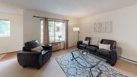 Located at 1718 E Lincoln Rd in Spokane, Washington, this single-floor residence offers a thoughtfully arranged layout that balances comfort and functionality. The living room is a welcoming space featuring plush carpeting, large windows that frame lush outdoor greenery, and ample natural light that brightens the neutral-toned walls. A leather seating arrangement and glass-top coffee table contribute to a clean, modern aesthetic.

The dining area flows seamlessly from the living room and is anchored by a glass dining table with upholstered chairs, positioned conveniently adjacent to the kitchen. The kitchen features warm wood cabinetry, a white appliance suite including a range and over-the-range microwave, tile backsplash, and durable wood-look flooring. A pass-through counter with bar seating connects the kitchen to the dining space, enhancing the open feel.

The bedroom is generously sized with soft carpeting, a large window offering pleasant outdoor views, a sliding-door closet providing ample storage, and a baseboard heating unit. The bathroom is well-appointed with wood-look flooring, a wide vanity with an extended mirror, and clean, neutral finishes throughout. Overall, the space presents a comfortable and practical living environment in a quiet Spokane neighborhood.