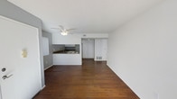 This single-floor space offers a clean, open-concept layout that seamlessly connects the living area and kitchen, creating a sense of spaciousness and ease of movement. Rich, dark hardwood flooring runs throughout, providing a warm contrast against the crisp white walls and ceilings. A ceiling fan centered in the main living area offers both functional air circulation and subtle visual interest overhead.

The kitchen is defined by a peninsula counter that separates it from the living space while maintaining an open feel. Dark countertops complement gray-toned lower cabinetry, while white upper cabinetry and a simple backsplash keep the aesthetic light and cohesive. A built-in range hood and under-cabinet appliances add to the kitchen's practical design.

Toward the back of the unit, a set of white built-in closet doors provides ample storage, and the entry door is neatly integrated into the same wall. A floor-level vent indicates a central HVAC system running throughout. Vertical blinds near the living area suggest a window or sliding door that can admit natural light. The overall ambiance is minimal and move-in ready, with a neutral palette that offers a versatile backdrop for a variety of interior styles.