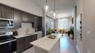 This single-story residence at Indigo WPB in West Palm Beach, Florida, offers a thoughtfully designed open-concept layout with contemporary finishes throughout. The kitchen serves as a central feature, equipped with dark charcoal shaker-style cabinetry, gold hardware accents, stainless steel appliances, a subway tile backsplash, and a generous quartz-topped island with pendant lighting above—ideal for both meal preparation and casual dining. Light wood-style flooring flows seamlessly from the kitchen into the living area, which benefits from large windows allowing ample natural light and views of the surrounding greenery. The living space provides a comfortable, modern atmosphere well-suited for relaxing or entertaining. The bedroom features a distinctive geometric accent wall, plush carpeting, and windows with sheer draping that invite natural light while maintaining privacy. The bathroom is finished with marble-style tile flooring, a dual-sink vanity with dark cabinetry and backlit mirrors, and direct access to a walk-in closet. Throughout the residence, elevated details such as recessed lighting, gold fixtures, and high ceilings contribute to a polished, contemporary aesthetic consistent with modern West Palm Beach living.