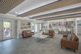 Located at 1056 Centerbrooke Ln in Suffolk, Virginia, this single-floor community amenity facility offers a thoughtfully designed and well-appointed gathering space for residents. The centerpiece of the space is a spacious lounge area, distinguished by its striking slatted wood ceiling panels with integrated ambient lighting that lend a warm, contemporary aesthetic throughout. Rich cognac leather sofas are arranged around a sleek marble-top coffee table, while a modern linear fireplace flanked by a stacked wood display adds a cozy focal point. Large French doors flood the lounge with natural light and provide views of the surrounding greenery outdoors. Adjacent to the lounge is a generously sized recreation and dining zone featuring a billiards table, multiple modern dining tables with yellow-accented seating, and open wall shelving — ideal for social gatherings. The fully equipped kitchen is outfitted with light wood cabinetry, stainless steel appliances, a subway tile backsplash, and a marble countertop island with pendant lighting overhead. A secondary kitchenette area with its own window and breakfast nook provides additional functionality. Polished large-format tile flooring flows seamlessly throughout, unifying the open, airy layout of the entire space.