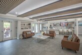 Located at 1056 Centerbrooke Ln in Suffolk, Virginia, this single-floor community amenity facility offers a thoughtfully designed and well-appointed gathering space for residents. The centerpiece of the space is a spacious lounge area, distinguished by its striking slatted wood ceiling panels with integrated ambient lighting that lend a warm, contemporary aesthetic throughout. Rich cognac leather sofas are arranged around a sleek marble-top coffee table, while a modern linear fireplace flanked by a stacked wood display adds a cozy focal point. Large French doors flood the lounge with natural light and provide views of the surrounding greenery outdoors. Adjacent to the lounge is a generously sized recreation and dining zone featuring a billiards table, multiple modern dining tables with yellow-accented seating, and open wall shelving — ideal for social gatherings. The fully equipped kitchen is outfitted with light wood cabinetry, stainless steel appliances, a subway tile backsplash, and a marble countertop island with pendant lighting overhead. A secondary kitchenette area with its own window and breakfast nook provides additional functionality. Polished large-format tile flooring flows seamlessly throughout, unifying the open, airy layout of the entire space.