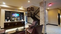This lobby presents a warm and inviting atmosphere through a thoughtful blend of rustic and contemporary design elements. A prominent exposed brick column serves as a striking architectural focal point, complementing the stone-accented wall behind the reception desk. The front desk area is finished in dark wood cabinetry with brass hardware, and features a built-in entertainment unit housing a flat-screen television alongside a beverage vending display. Recessed lighting throughout the space provides a well-lit yet comfortable ambiance, accentuating the warm earth tones of the walls and finishes. A seating area furnished with rich burgundy leather armchairs and a glass-top coffee table offers a comfortable waiting space for guests. A dark-railed staircase with coordinating wood finishes leads to the upper level, adding visual depth to the space. The flooring features a combination of tile with a decorative inlay pattern, enhancing the overall character of the lobby. Together, these elements create a cohesive, welcoming environment with a distinctly upscale yet approachable aesthetic.