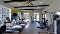 This fitness center is designed to accommodate a comprehensive workout experience within a single, open-plan floor. The space is defined by its bright, airy atmosphere, achieved through multiple large windows that line the walls and invite generous amounts of natural light, offering views of the outdoors. Recessed ceiling lights and two ceiling fans ensure comfortable conditions throughout the room.

The interior design features a clean, modern aesthetic with white walls accented by bold yellow and blue color blocks, adding visual energy to the environment. Dark exposed ceiling beams provide a subtle architectural contrast against the light-colored ceiling.

The cardio zone is well-stocked with a row of TRUE-brand treadmills and additional cardio machines, all positioned to take advantage of the natural light and outdoor views. On the opposite side of the space, strength and resistance training equipment is available, including a cable machine and weight bench. A dedicated functional training station houses medicine balls, resistance tools, and stability balls neatly organized on a rack. A wall-mounted flat-screen television is also present, adding entertainment options for users during their workouts. The flooring is a durable, neutral-toned gym-appropriate surface throughout.