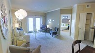 This single-floor residence offers a thoughtfully designed layout that balances comfort and style. The living area serves as the heart of the space, featuring plush wall-to-wall carpeting, elegant crown molding, and soft neutral tones accented by sage green walls that create a calming ambiance. A set of French doors floods the room with natural light while providing access to an outdoor area, enhancing the sense of openness and airiness. The living space is generously proportioned, providing ample room for multiple seating arrangements, making it well-suited for both relaxing and entertaining. Decorative circular wall accents add a modern, artistic touch to the interior design. An open doorway connects the living area directly to the bedroom, which continues the warm, neutral color palette and is finished with coordinating soft furnishings in blue and green tones. The bedroom benefits from its own lighting and framed wall art, contributing to a cozy yet refined atmosphere. Overall, the space presents a cohesive design aesthetic with consistent flooring, trim details, and a harmonious color scheme that flows naturally from room to room, creating a welcoming and well-organized living environment.